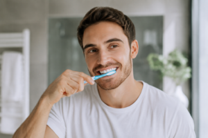 a man wearing a white tshirt with brown hair brushing his teeth in the mirror