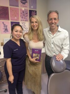 a photo of tracy tang and dr james goolnik with a patient, holding James' kick sugar cookbook in the dental surgery